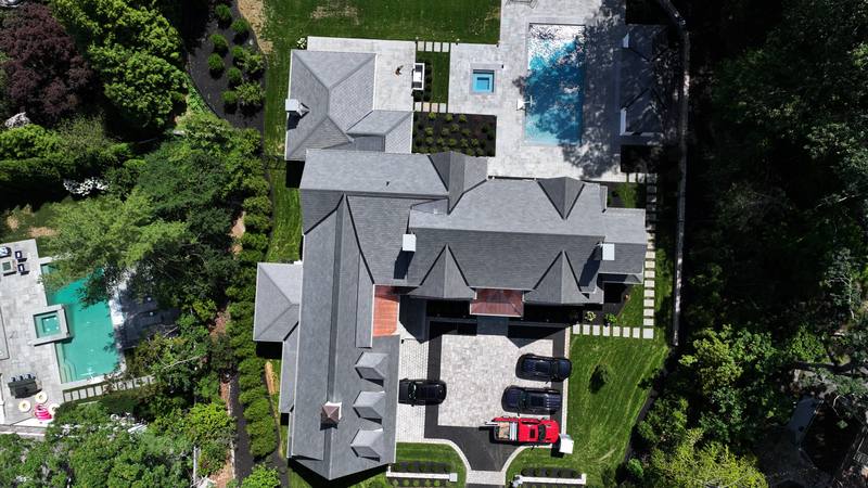 Large home with a gray shingle roof beside a pool and courtyard, aerial view